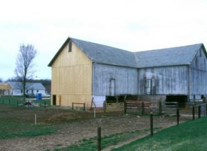 Two-tone barn under renovation in a rural field, with a new light wood left side and weathered right side and a fence in the foreground.