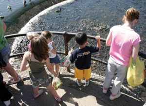 Children stand at a railing overlooking a rocky water area, some holding bags and looking down. Safer, sunny day at a river or harbor railing scene.