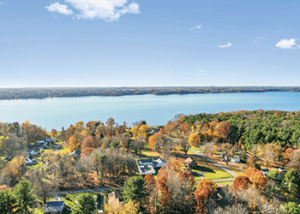 Aerial view of a lake with autumn trees and a small neighborhood at the shore under a clear blue sky.