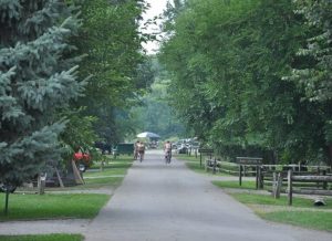 Park path lined with trees with cyclists riding in the distance; tents and an umbrella visible beyond.