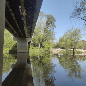 Under-bridge view of a calm river with green trees reflected in the water, the bridge spans from the left.