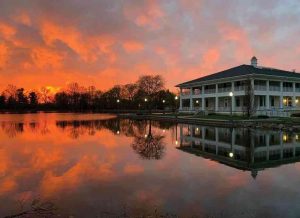 Sunset over a calm lake reflecting a white two-story pavilion with lights on by the shore.