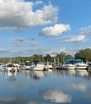 Boats docked at a marina on calm water with reflections and a blue sky full of clouds.