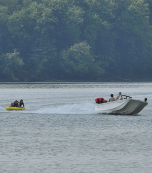 Speedboat tows a yellow inflatable raft carrying people across a calm lake, with a forested shoreline in the background.