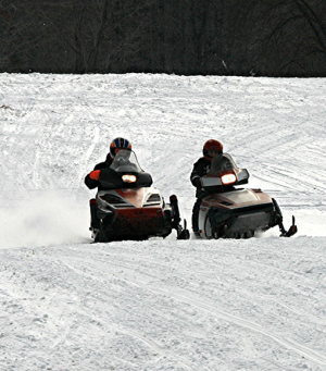 Two people on snowmobiles riding side by side on a snow-covered trail, kicking up powder behind them.