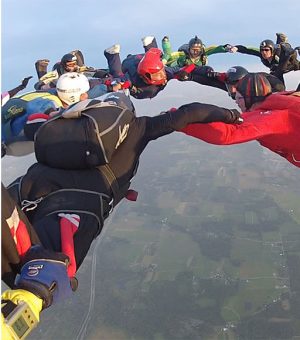 A group of skydivers forming a circle in freefall, holding hands mid-air with colorful suits and helmets above a rural landscape.