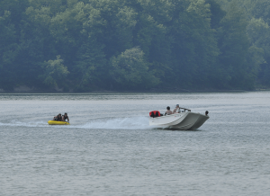 A white motorboat tows a yellow inflatable raft with three people across a calm lake; tree-covered shoreline in the distance.