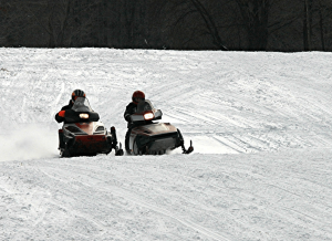 Two people on orange snowmobiles riding side by side down a snowy slope with a forest in the background, winter gear and helmets visible in the foreground.