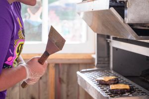 Cook in a purple shirt wearing gloves flips a burger on a stainless grill with cheese slices on top patty pieces visible inside the grate.