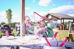 Children blowing colorful bubbles at an outdoor bubble station, with cups and pink wands on a table under a blue sky.