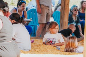 Group of adults and children gathered outdoors around a snack table, with blue chairs and a wooden post in the background.