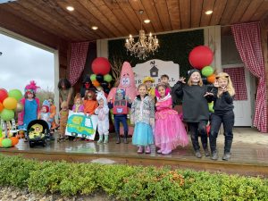 Group of children in colorful costumes on a wooden stage, posing for a photo with balloons and a Halloween backdrop.“ (stage with red-checked curtains)