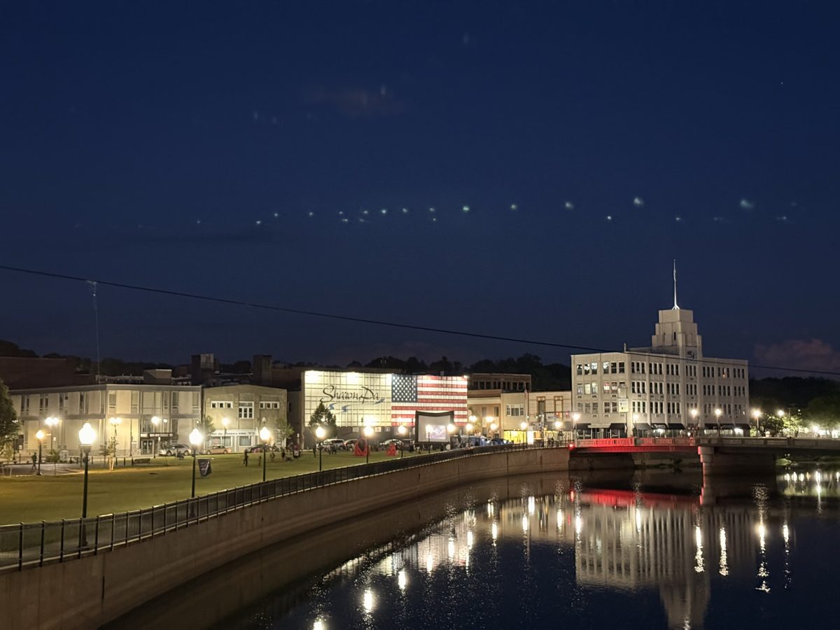 Night view of a city waterfront with illuminated buildings, a red-lit bridge, and reflections on the water surface at dusk.