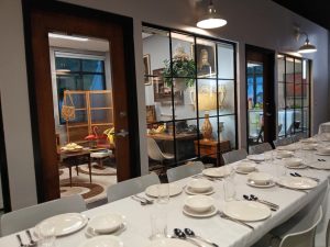 Long white banquet table set with plates, bowls, and cutlery for a formal dinner inside a showroom, seen through glass partition walls.