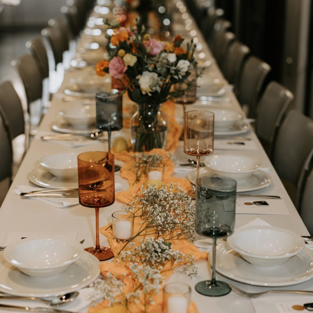 Long banquet table set for a formal event with orange runners and colorful floral centerpieces.