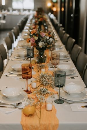 Long banquet table set for a formal event with orange runners and colorful floral centerpieces.