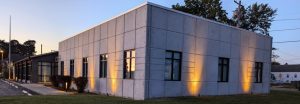 Modern concrete building at dusk with warm ground lights along the wall and a sidewalk in front.